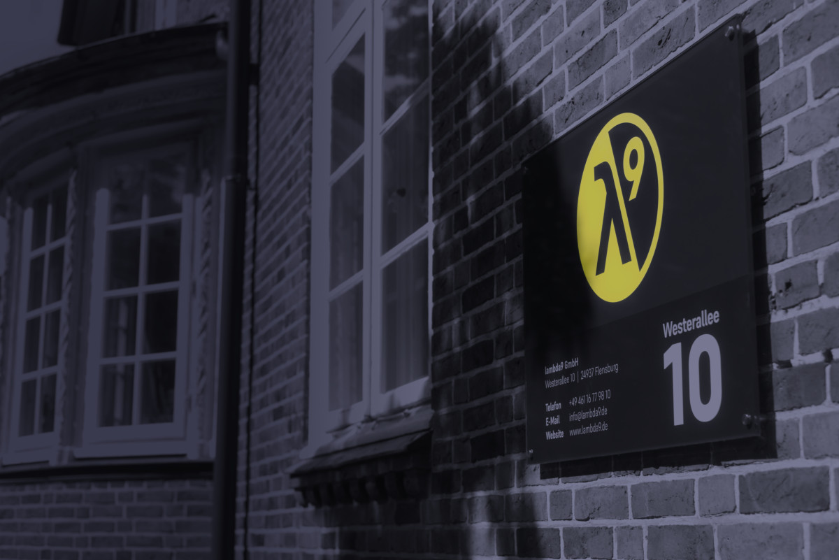 Vor einer Häuserwand ist ein Schild mit Logo und Adresse der Firma lambda9 montiert.